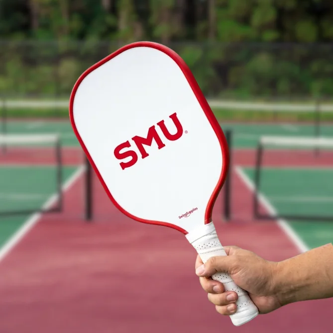 A hand holding a white pickleball paddle printed with the SMU wordmark over a court